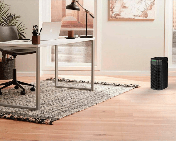Home office with a white desk, laptop, and a black tower air purifier on the hardwood floor by the rug.