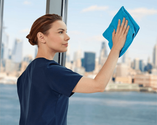 Woman in a navy shirt wipes a window with a bright blue cloth, with a city skyline visible outside.