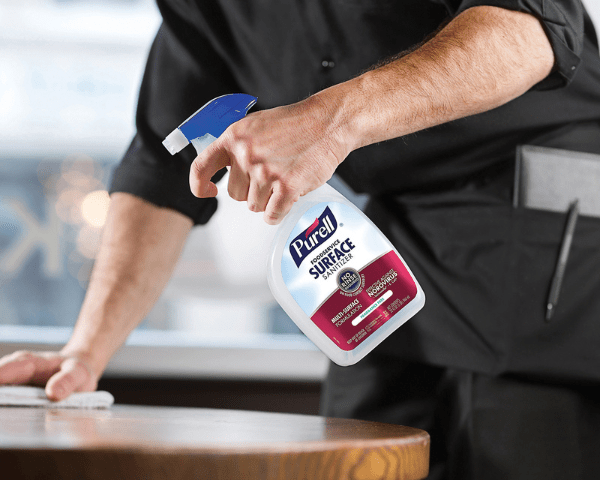 Person wearing black shirt uses a spray bottle to sanitize a wooden table while wiping it with a cloth in a cleaning action.