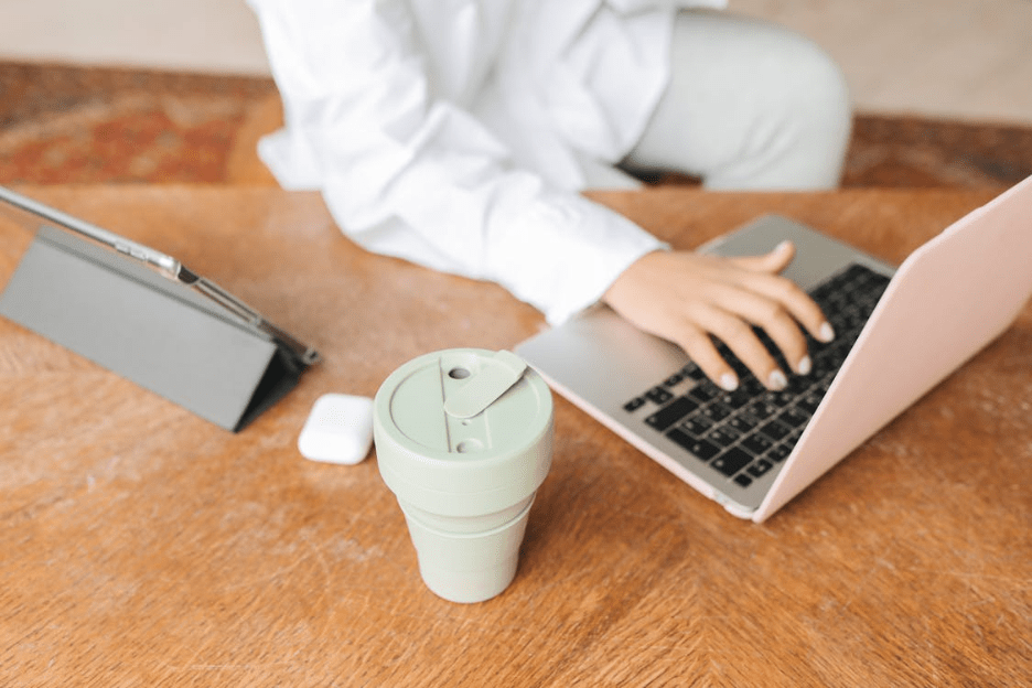 A person working remotely on a laptop with a tablet and reusable cup on a wooden table. Photographer: Polina Tankilevitch