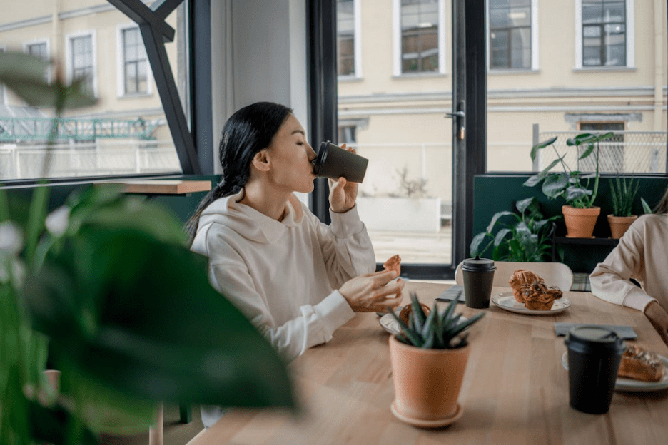 An Asian woman sipping coffee, seated indoors at a cozy cafe with snacks and plants. Photographer: Ron Lach