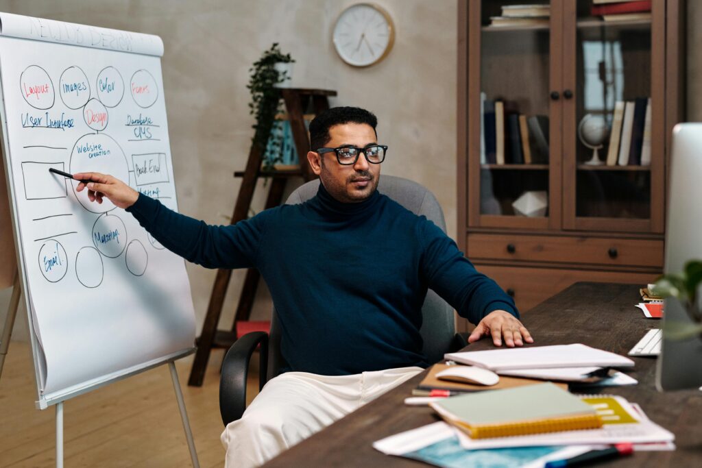 Man teaching online, pointing at a flipchart in a cozy office space. Perfect for online education themes. Photographer: Diva Plavalaguna
