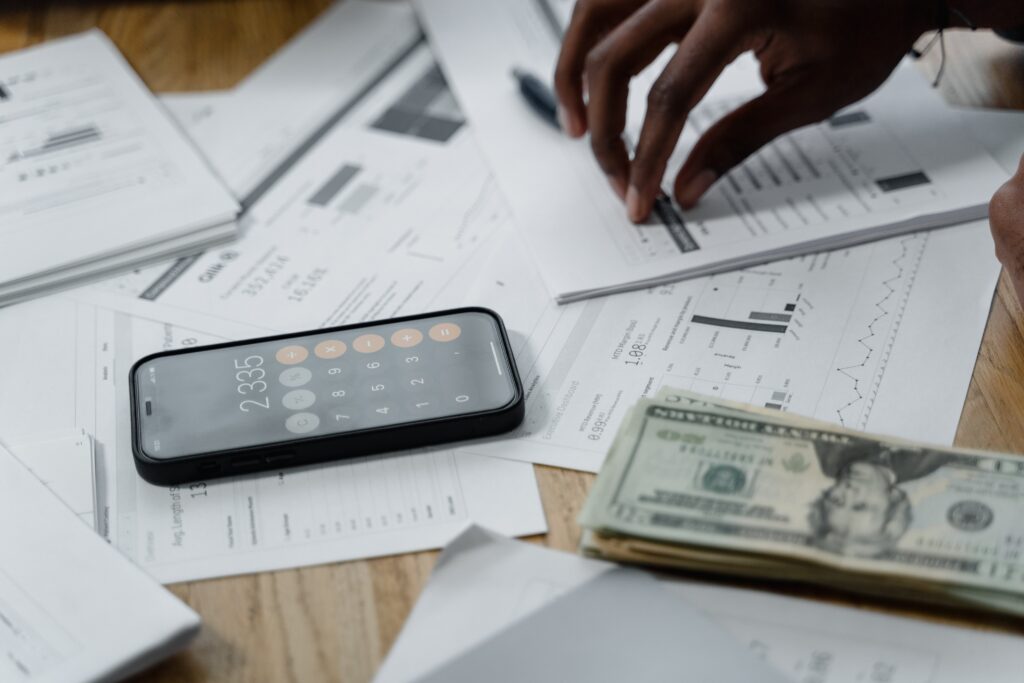 Smartphone showing a calculator app on a cluttered desk with financial documents, charts, and a hand grabbing a page for data. Photographer: Tima Miroshnichenko